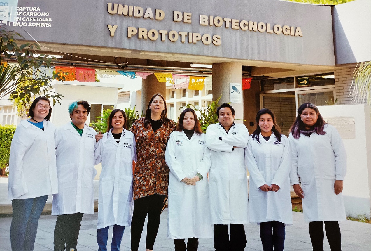 8 people stood in a row smiling. Seven are wearing white lab coats with UNAM written on them. Above them on a building is written Unidad de Biotecnologia y prototipos